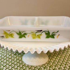 May include: A white divided casserole dish with a hand-painted ivy design in green and yellow, resting on a white, ruffled-edge cake stand. The dish has two handles and is perfect for serving. A decorative piece for the kitchen.