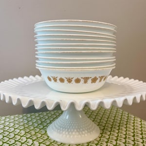 Peut inclure: Une pile de bols blancs avec un motif floral doré sur un support à gâteau blanc à bord festonné. Le support est sur une nappe à motifs verts et blancs.