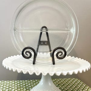 May include: A clear glass divided serving dish with a ridged pattern, displayed on a white, ruffled-edge cake stand. The dish is supported by a black metal stand with decorative scrollwork. The cake stand has a pedestal base.