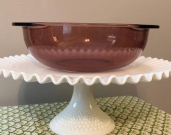 May include: A translucent plum-colored glass bowl with side handles resting on a white milk glass pedestal stand. The stand features a scalloped edge and a textured hobnail pattern on the base, providing a stable elevated display.