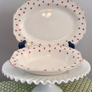 May include: An oval serving platter and a matching shallow bowl made of white ceramic, both featuring a repeating pattern of small red flowers and green stems. Each piece has a scalloped edge with a thin gold trim along the outer rim.