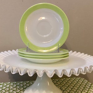 May include: A set of four white and green vintage saucers with a scalloped edge. The saucers are stacked on a white cake stand with a scalloped edge.