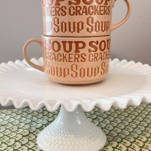 May include: Two stacked peach-colored soup mugs with handles, each printed with "SOUP CRACKERS Soup Soup" in brown lettering. The mugs sit on a white, ruffled-edge cake stand, atop a green and white patterned tablecloth.