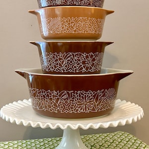 May include: A stack of four vintage casserole dishes in shades of brown and tan, each adorned with a white floral design. The dishes are displayed on a white, scalloped-edge cake stand, creating a charming, retro aesthetic.