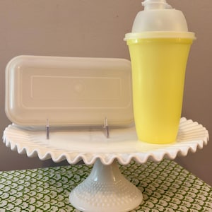 May include: A white milk glass cake stand with a ruffled edge and hobnail base. Resting on the stand is a rectangular white plastic lid and a tall, yellow plastic shaker cup with a translucent white lid featuring a flip-top spout closure.