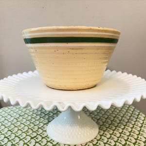 May include: A vintage beige mixing bowl with a dark green stripe, resting on a white, ruffled-edge cake stand. The bowl has a textured surface and a classic design, perfect for kitchen decor or baking.