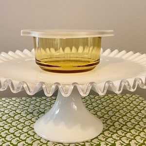 May include: A white cake stand with a scalloped edge and a pedestal base. A small amber glass bowl with a white lid sits on top of the stand.