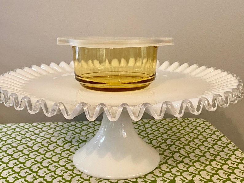 May include: A white cake stand with a scalloped edge and a pedestal base. A small amber glass bowl with a white lid sits on top of the stand.