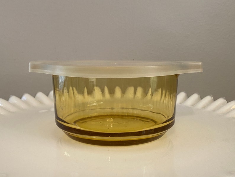 May include: A small amber glass bowl with a white plastic lid. The bowl has a textured design and is perfect for storing small items.