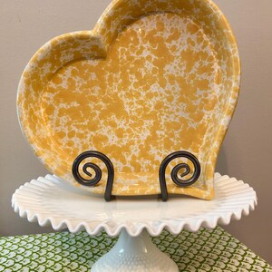 May include: A heart-shaped ceramic dish featuring a yellow and white speckled pattern. The dish is held upright by a black metal scrollwork stand, which rests on a white milk glass pedestal cake stand with a ruffled edge and a hobnail base.