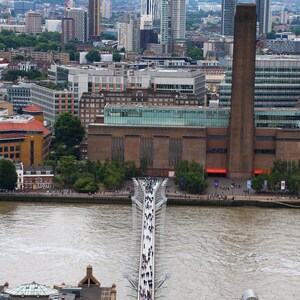 Puede incluir: Vista aérea de un puente peatonal sobre un río en una ciudad. El puente está hecho de metal y tiene una superficie blanca. La gente camina por el puente. En el fondo, hay edificios y árboles.