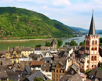 May include: A view of a small town in Germany with a church steeple in the foreground. The town is situated on a river with a green hill in the background.