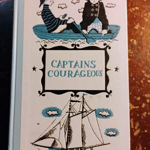 May include: A vintage book cover with the title "Captains Courageous." The cover features illustrations of two figures in a boat, a sailboat, and the title in a decorative frame. The color palette includes blue, black, and white.