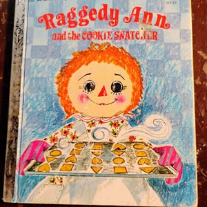 May include: A vintage "Little Golden Book" titled "Raggedy Ann and the Cookie Snatcher." The cover features a Raggedy Ann illustration holding a tray of cookies. The background is light blue with a checkered pattern.