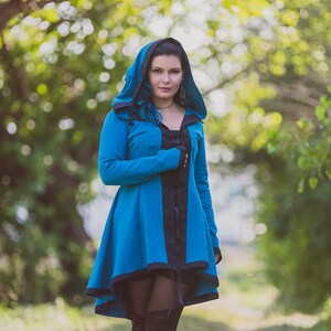 Può includere: Una donna indossa una giacca blu con cappuccio, bordi neri e una chiusura con lacci neri sul davanti. È in piedi in un ambiente boschivo con alberi verdi e un sentiero di terra.