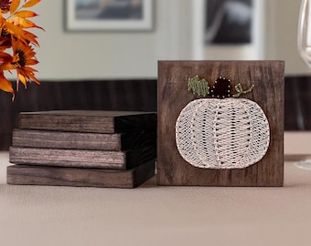 May include: A set of square dark brown wooden coasters. One coaster stands upright, displaying a string art design of a pumpkin created with light-colored thread, a dark brown stem, and green leaves. The other four coasters are stacked horizontally.