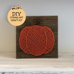 May include: A square dark brown wooden board featuring a pumpkin design created with orange string wrapped around small metal nails. A small brown string stem sits atop the pumpkin. A circular label reads mini DIY STRING ART CRAFT KIT.