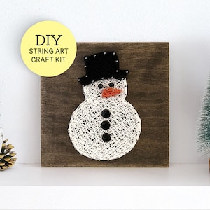 May include: DIY string art craft kit featuring a snowman design. The snowman is crafted with white string on a dark wooden square, with a black hat, orange nose, and black button eyes. A yellow circle with text "DIY STRING ART CRAFT KIT" is in the upper left.