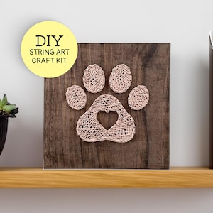May include: A square dark brown wooden board featuring a string art design of a paw print with a heart-shaped center. A yellow circular label in the upper left corner displays the black text DIY STRING ART CRAFT KIT in capital letters.