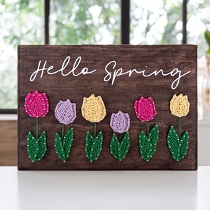 May include: A wooden sign with the text "Hello Spring" in white paint. The sign is decorated with six colorful string art tulips in shades of pink, purple, and yellow.