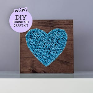 May include: A square dark brown wooden board featuring a heart shape created with bright blue string wrapped around silver nails. A circular light purple label in the upper left corner displays the text mini DIY STRING ART CRAFT KIT.