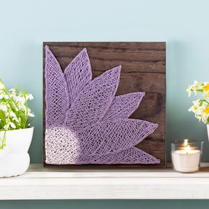 May include: A handcrafted string art piece featuring a purple flower design on a wooden square. The artwork is displayed on a white shelf, with white flowers in a white pot on the left and yellow flowers in a pink pot on the right.