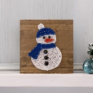 May include: A square wooden plaque featuring a string art snowman. The snowman is created with white string, wearing a blue string hat and scarf, an orange string nose, and three black buttons for the body. The design is mounted on a brown board.