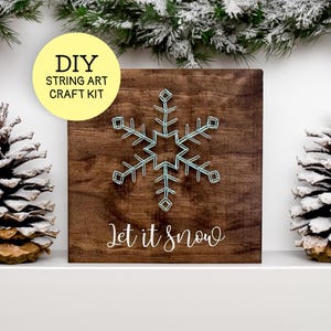 May include: A DIY string art craft kit featuring a snowflake design on a wooden board. The board is brown, and the snowflake is made with light blue string. The text "Let it snow" is written in white script below the snowflake. Two snow-covered pinecones flank the board.