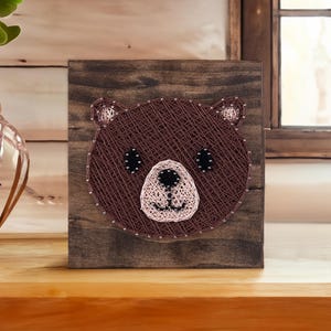 May include: A brown wooden frame with a string art design of a bear's face. The bear's nose and mouth are pink, and its eyes are black.