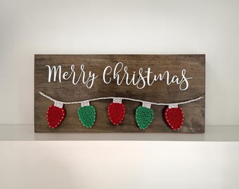 May include: A wooden sign with the text "Merry Christmas" in white lettering. A string of red and green string lights is strung across the bottom of the sign.