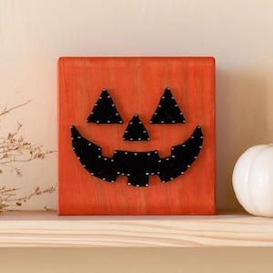 May include: A square orange wooden block featuring a jack-o'-lantern face created with black string art. The design includes two triangular eyes, a small triangular nose, and a wide, jagged, toothy grin, all formed by black thread wrapped around nails.