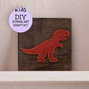 May include: A DIY string art craft kit for kids featuring a red Tyrannosaurus Rex design on a dark brown wooden square. The kit includes the text "kids DIY STRING ART CRAFT KIT" on a purple circular sticker.