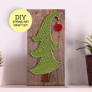 May include: A DIY string art craft kit featuring a green string Christmas tree on a rectangular wooden board. The tree has a brown trunk and a small red circular ornament hanging from the top branch. The text DIY STRING ART CRAFT KIT is visible.