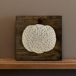 May include: A square dark brown wooden plaque featuring a string art pumpkin design. The pumpkin is created with white string wrapped around silver nails, forming a detailed outline and interior pattern, topped with a small dark brown stem.