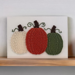 May include: A rectangular white board featuring three pumpkins created with string art. The pumpkins are colored cream, orange-red, and dark green, each topped with a dark brown stem and thin, curly green wire tendrils extending to the sides.