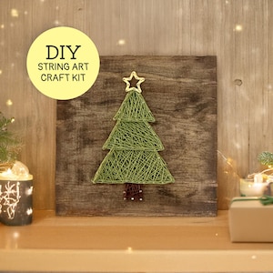 May include: A DIY string art craft kit featuring a green string Christmas tree on a dark brown wooden board. The tree is topped with a gold star outline and has a brown string trunk. A yellow circular label displays the text DIY STRING ART CRAFT KIT.