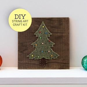 May include: DIY string art craft kit featuring a Christmas tree design. The tree is made of green string on a dark brown wooden square, adorned with colorful string lights. A red and a green glittery ornament are on either side.