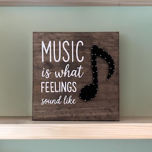 May include: A square dark brown wooden sign featuring the white text MUSIC is what FEELINGS sound like and a black string art musical note. The design is created with white nails outlining the note shape on the stained wood surface.