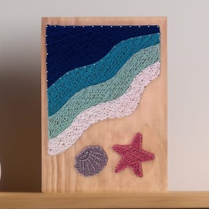 May include: A wooden string art piece depicting a beach scene. The artwork features layered waves in shades of blue, teal, and white. A red starfish and a gray seashell are also included. The art is mounted on a light-colored wooden board.