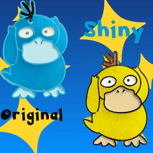 May include: Two flat character ornaments featuring a duck-like creature. One is bright blue with a light blue beak and black hair, labeled Shiny. The other is yellow with a beige beak and brown hair, labeled Original, set against a blue background.
