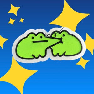 May include: A lime green, two-frog shaped ornament with a black outline. One frog has a long, pointed snout touching the other frog. The ornament has a white border and is set against a blue background with yellow four-pointed star shapes.