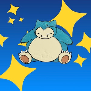 May include: A blue and cream-colored enamel pin shaped like a sleeping creature with a large round belly, small ears, and clawed feet. The design features a dark blue background with several yellow four-pointed star shapes surrounding the character.
