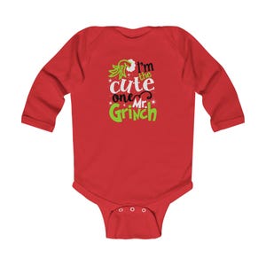 May include: A long-sleeve red baby bodysuit featuring a graphic print in green, white, and black. The text reads I'm the cute one Mr. Grinch, accompanied by a stylized green hand illustration and small star accents centered on the chest area.