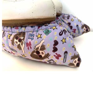 May include: Purple shoe covers with a pattern of girls wearing sunglasses, stars, hearts, and other accessories. The shoe covers are made of a soft, stretchy fabric and are designed to protect shoes from dirt and grime.