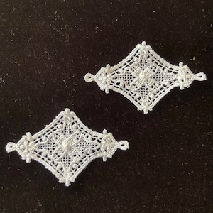 Op de afbeelding: Twee witte, ruitvormige kanten applicaties. Elke applicatie heeft een gedetailleerd bloemmotief in het midden, omgeven door een opengewerkt patroon. De randen zijn afgewerkt met kleine lussen. De applicaties worden op een donkere achtergrond weergegeven.