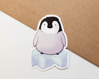 以下が含まれることがあります： 黒い頭と白い顔の漫画のペンギンのステッカー。水色の氷山の上に立っています。ペンギンの体は薄紫色です。ステッカーは白い表面にあり、背景には茶色の封筒があります。