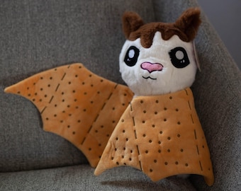 Könnte beinhalten: Ein Plüsch-Fledermaus-Spielzeug mit braunem Fell und einem weißen Gesicht. Die Fledermaus trägt eine braune und beige Decke mit einem Tupfenmuster. Die Fledermaus hat große schwarze Augen und eine rosa Nase.