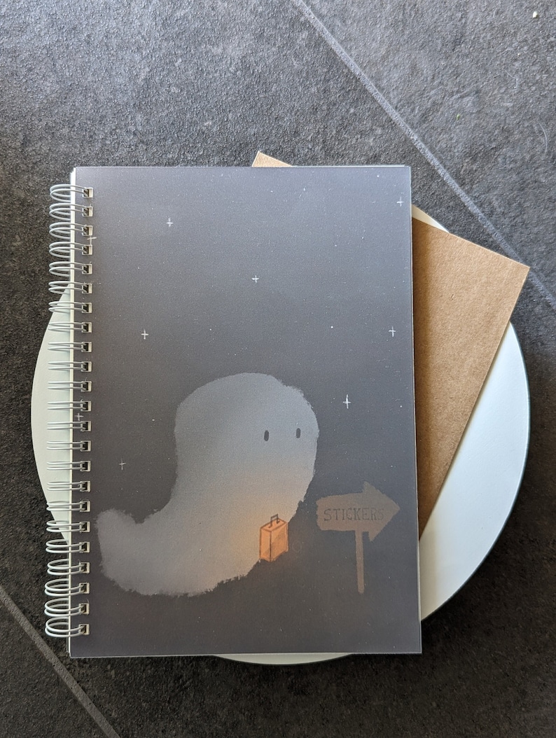 May include: A gray spiral bound notebook with a ghost illustration. The ghost is holding a small orange suitcase and there is a sign that says "Stickers" with an arrow pointing to the right. The background is a dark blue with white stars.