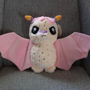 May include: A plush bat toy with a cream-colored body, pink wings, and a pink and yellow head. The bat has large black eyes, a purple nose, and small colorful sprinkles on its body. It is sitting on a gray chair.