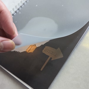 May include: A black spiral bound notebook with a clear plastic cover. The cover has a white cloud and stars design. The notebook has a black and white illustration of a sign that says "Stickers" with an arrow pointing to the right. The illustration also includes a small brown box.